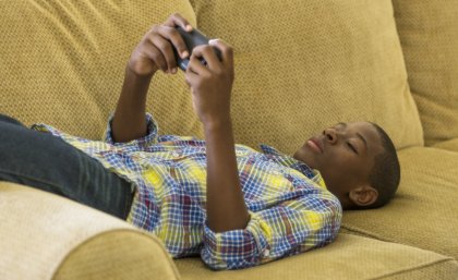 A boy lays on a couch watching something on a hand-held screen device.